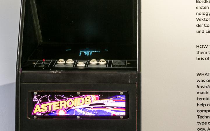 Ein »Asteroids« Arcade Automat vor einer Wand mit einem Beschreibungstext. 