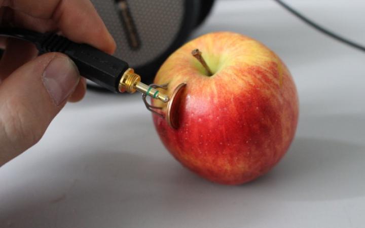 In einem Apfel stecken eine Kupfermünze und ein Zinkdraht, jemand hält an beide metallenen Gegenstände einen Klinkenstecker durch den nun Strom fließt.