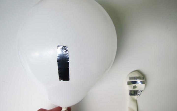Ein aufgeblasener weißer Ballon mit einem Streifen glänzenden metallenen Klebeband auf weißem Grund. Daneben ein noch nicht aufgeblasener Ballon mir vielen solcher Klebestreifen.