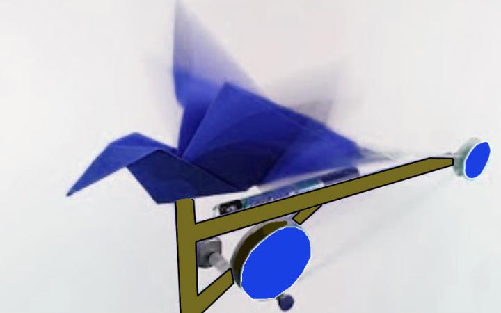 Ein aus blauem Papier gefalteter Origami-Vogel ist auf eine Unterkonstruktion mit Rädern geklebt. Außerdem ist eine Batterie an dem Objekt angeschlossen.