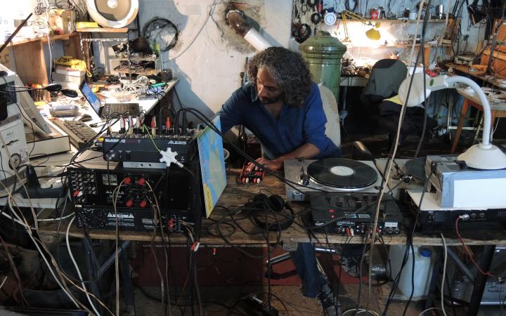 Zu sehen ist ein Mann in einem Raum voll Kabel und Geräte, der Musik produziert. 