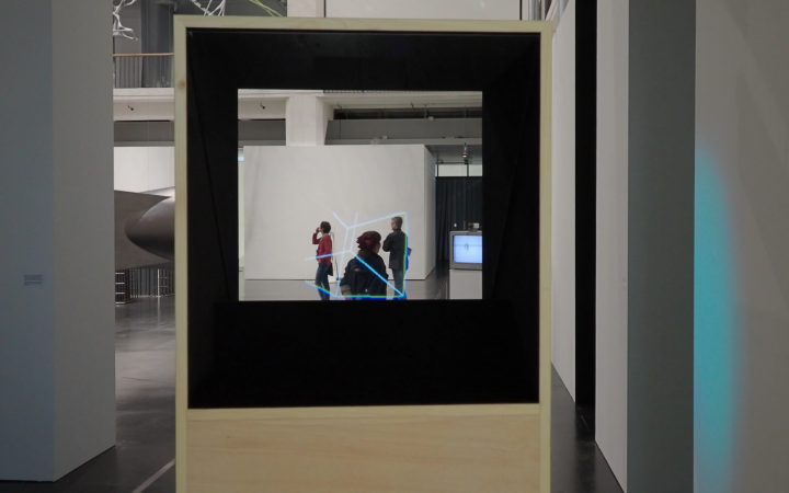 Blick durch einen Kasten mit einer durchsichtigen Platte in einen Raum. Im Raum ist eine animierte geometrische Figur zu sehen.