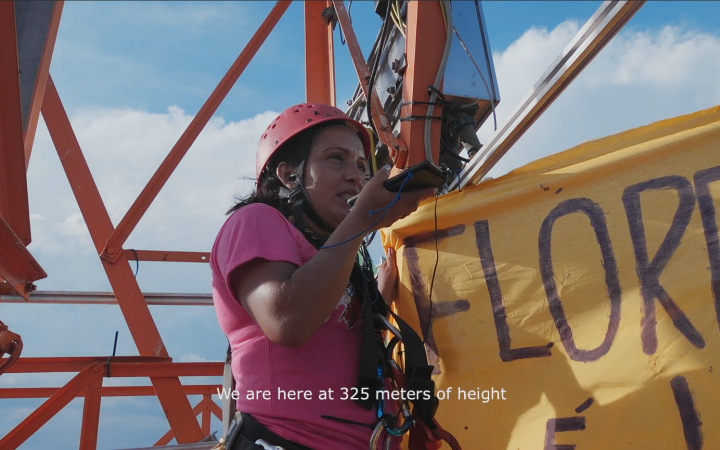 The picture shows a woman on a steel frame holding a large banner in one hand and a smartphone in the other, which she talks into. You can read: we are at 325 meters of height. 
