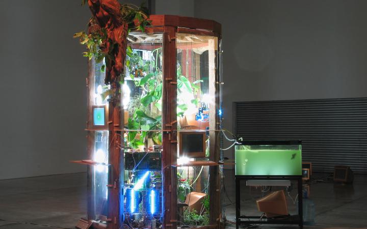 An einer Art Gewächshaus, das mit Strahlern von innen beleuchtet wird, befinden sich Planzen und Computer. Daneben ist ein Aquarium mit Schläuchen angeschlossen. Am oberen Rand des Glaskastens befindet sich eine hölzerne Galionsfigur.