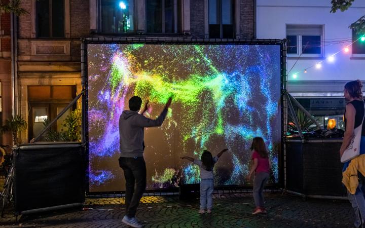 In the pedestrian zone there is a screen with brightly colored particles, and adults and children move in front of it.