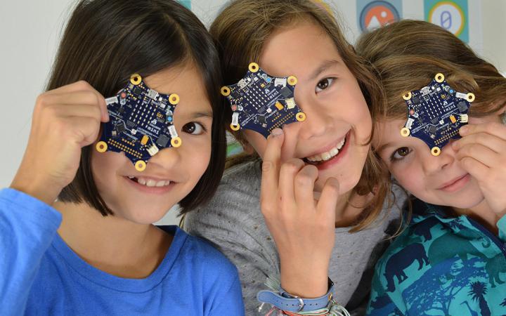Photo of three young girls each holding a Calliope mini circuit board in front of their face.