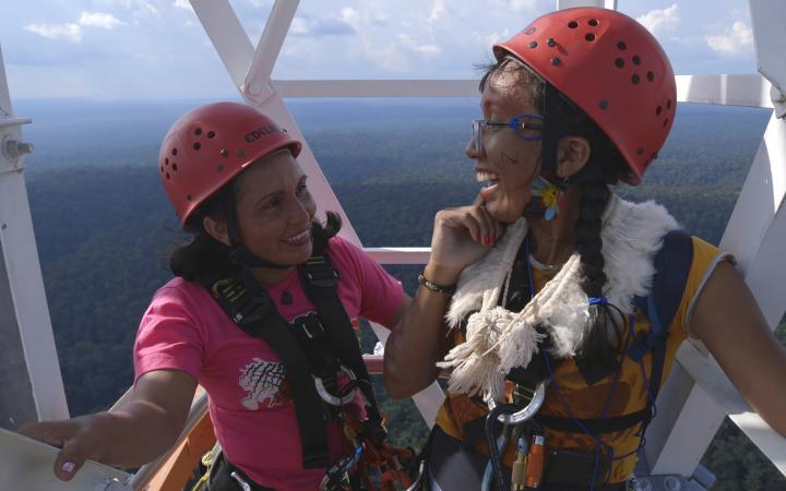 Two women can be seen standing high up on a tower. They wear safety helmets and laugh. In the background you can see forest up to the horizon.