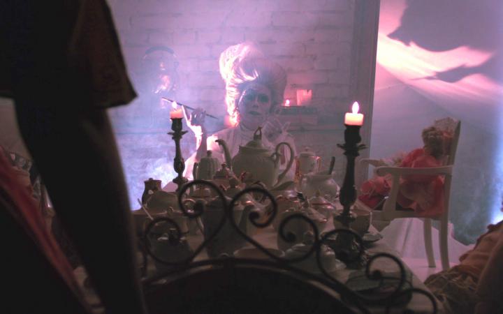 A smoking person is sitting in a room with stone walls. She is wearing a wig from the 18th century, is heavily made up and sits at a table with a long candle burning on it. Around the table are dolls.