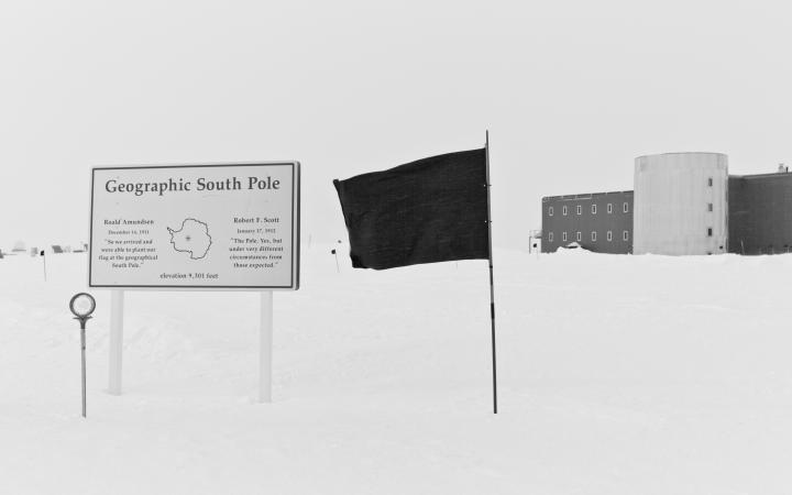 Das Bild zeit eine schwarze Flagge im Schnee neben einer Infotafel über den Südpol und einem Haus, was im Hintergrund zu sehen ist.