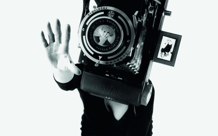 The black and white photo shows a woman in a black mini dress on her knees. Her face is covered by an oversized camera. 