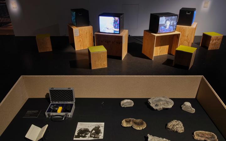 Foto aus der Ausstellung »Critical Zones« im Hintergrund laufen Videos auf Röhrenbildschirmen, im Vordergrund liegen Fotos und Gesteinsproben.