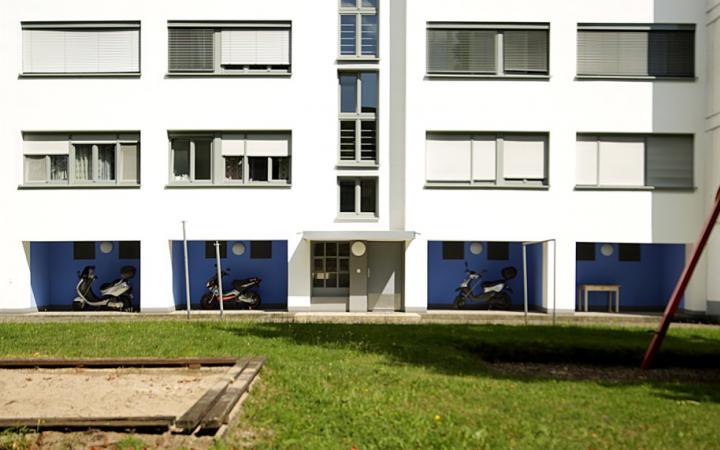 Zu sehen ist ein großes Weiße Gebäude mit einer Galerie im Erdgeschoss, deren Wände blau gestrichen sind. Das Gebäude befindet sich in der Dammerstocksiedlung in Karlsruhe, die aus der Bauhaus Zeit stammt.