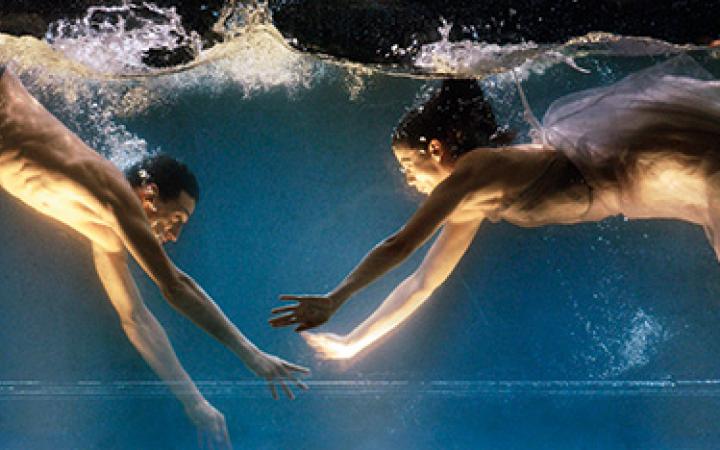 Ausschnitt aus Sasha Waltz "Dido & Aeneas". Zwei halbnackte Tänzer zeigen eine Choreografie in einem bis zum Bildrand gefüllten Wassertank, welcher von unten beleuchtet wird.