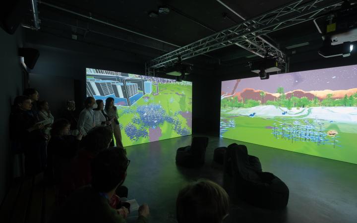 Hier sieht man das Werk »DO AIS DREAM OF CLIMATE CHAOS - SYMBIOTIC AI«. Zu sehen ist ein abgedunkelter Raum, indem sich mehrere Personen befinden. Im Hintergrund befinden sich zwei große Bildschirme, die animierte Landschaften abbilden.