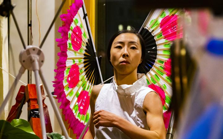 The photo shows the upper body and head of a Korean performer wearing two pink/light green coloured wooden fans as wings.