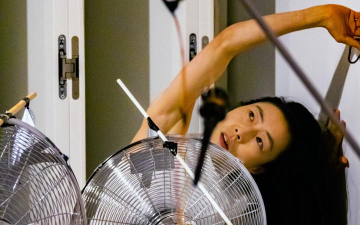 The photo shows a Korean performer bent to the left stretching her arm over her head while a large silver fan is standing in front of her.