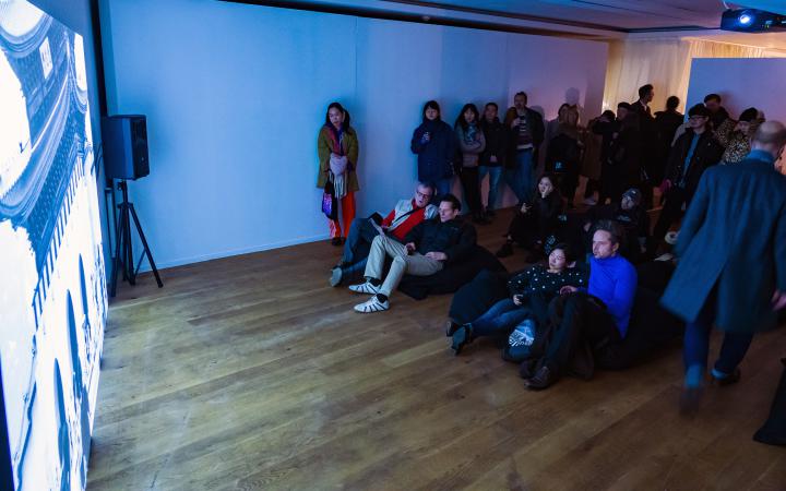 Zu sehen sind viele Menschen die vor einer großen Kunstprojektion stehen oder liegen. Der Raum hat einen Holzboden und wird von der Projektion blau erleuchtet.