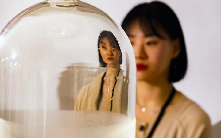 Das Foto zeigt eine Glasglocke hinter der eine koreanische Frau steht. Ihr Gesicht und Oberkörper reflektieren verzerrt in der Glasglocke.