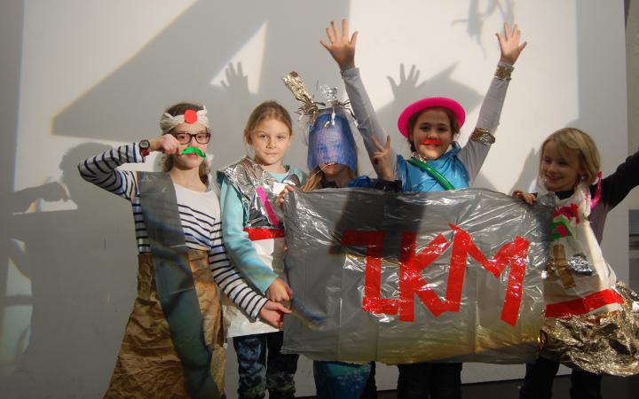 A group of girls dressed in clothes out of ordinary items are holding a banner, that has ZKM written on it with red tape.