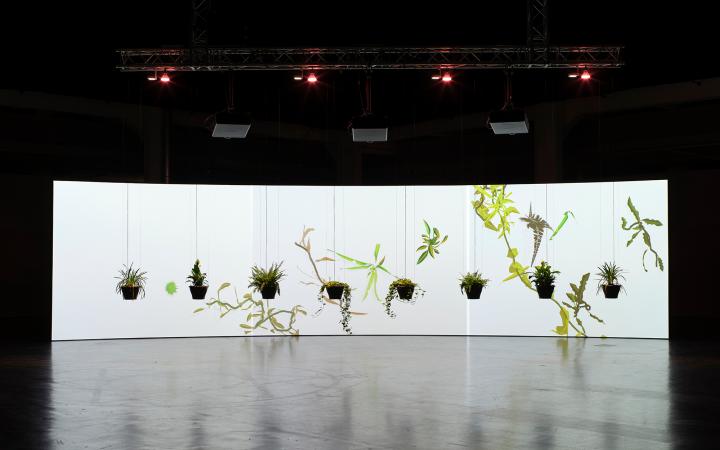 The frontal view of the artwork "Eau de Jardin" is shown. In the foreground, some plants hang in a semicircle from the ceiling and in the background a large canvas can be seen on which there are floral shapes. 
