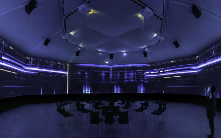 David Monacchi can be seen in a blue room. Sound tracks are projected on the walls. Chairs are placed in the center of the room.