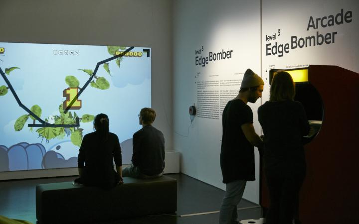 Ausstellungsansicht »EdgeBomber« mit Spielern vor einer Wandprojektion und einem Spieleautomat