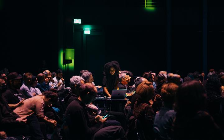 Jessica Ekomane während einer Performance an ihrem Laptop stehend zwischen einer sitzenden Menschenmenge