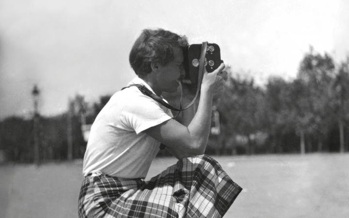 The black and white photograph shows a woman in squatting position with a camera.