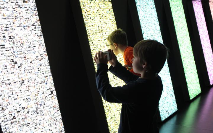 Zwei Kinder stehen vor einer Leuchtwand. Eines fotografiert gerade die Installation, das andere hat das Gesicht ganz nah am Kunstwerk um die kleinen Bilder genauer zu betrachten, die erleuchtet werden.
