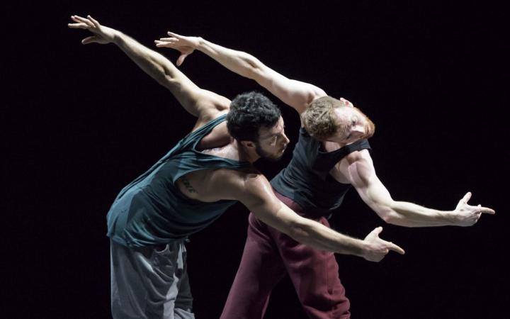 Two dancers in the choreography »Friends of Forsythe«. They stand together on a black stage and artfully contort their bodies parallel to each other.