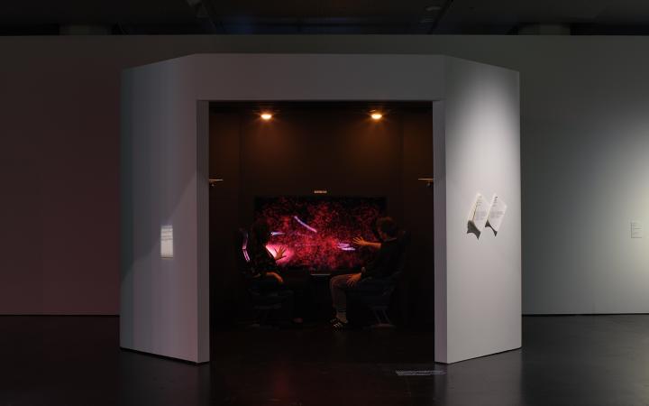 On display is the work "Haze Express". The picture shows two people looking at the work from one side each. It is the total view to see. The people are sitting in a small room where the work is and on a canvas you can see abstract shapes in red colors.