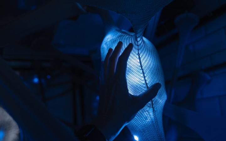 Hier sieht man das Werk »Human Bacteria Interfaces«. Eine Hand berührt ein blau-leuchtendes Objekt, das von einem Netz überzogen ist.