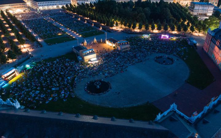 The photo shows an overview of the visitors to the premiere of the Schlosslichtspiele 2019. The photo was taken with a drone above the castle and shows a large crowd of people on the green meadow in front of Karlsruhe Castle.