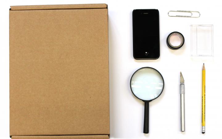 Zu sehen ist eine Lupe, ein Smartphone, eine Pappkiste, ein Cutter, ein Stift, eine Büroklammer, eine Kassettenhülle und Klebeband.