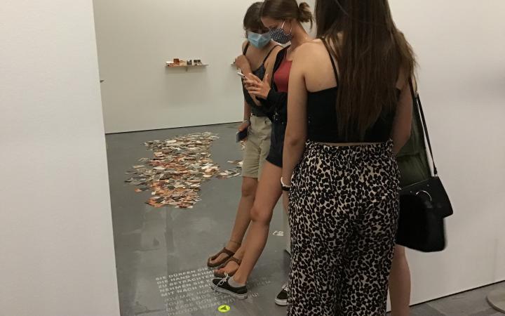 Zu sehen sind drei Mädchen, die ihre Füße auf einen Text auf dem Boden gestellt haben und davon ein Foto machen.
