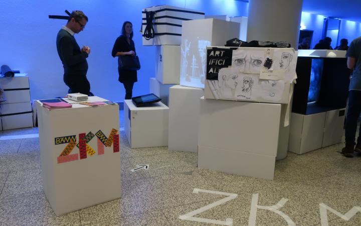 On display is a room with people and various drawings on stacked cardboard boxes as part of an event organised by the cultural academy.