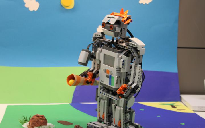 Vor einer bunten Papierkulisse, die Rasen eine Sonne und Wolken andeutet, steht ein Legoroboter.