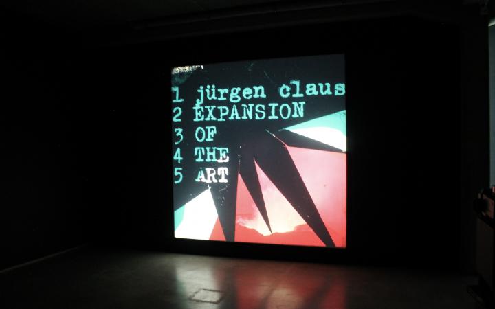 Photo of a multimedia projection, an image in pink, black and turquoise with geometric shapes. On it the text: »Jürgen Claus, Expansion of the Art«
