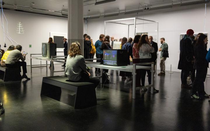 Exhibition view »Matter. Non-Matter. Anti-Matter« at ZKM | Center for Art and Media Karlsruhe, 2022. A person wears headphones and sits in front of a tube TV. In the background there is a crowd of people.