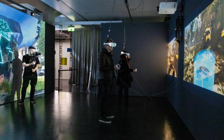 Ausstellungsansicht »Matter. Non-Matter. Anti-Matter« im ZKM | Zentrum für Kunst und Medien Karlsruhe, 2022. Auf der linken und rechten Seite sind zwei große Leinwände zu sehen. Zwei Personen tragen eine VR-Brille. 