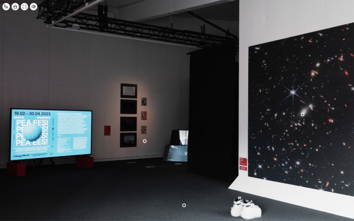 Blick in einen Ausstellungsraum, mit einem großen Bildschirm mit Informationstext auf der linken Seite, daneben mehrere Gemälde und Fotografien an der Wand. Auf der rechten Seite des Ausstellungsraum ist ein großes Bild mit funkelnden Sternen im schwarzen