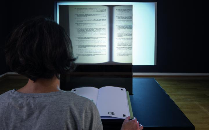 You can see the back of a person's head, and in front of it is an open book. On the wall opposite is a projection of an opened book.