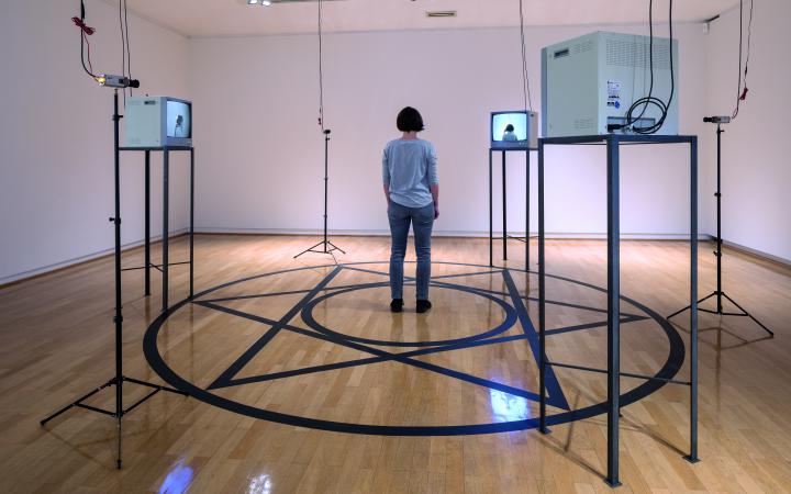 A room can be seen in which four monitors stand on a rack. A circle is glued to the floor, in the middle of which is a star and another circle. In the middle is a woman with her back to the viewer.