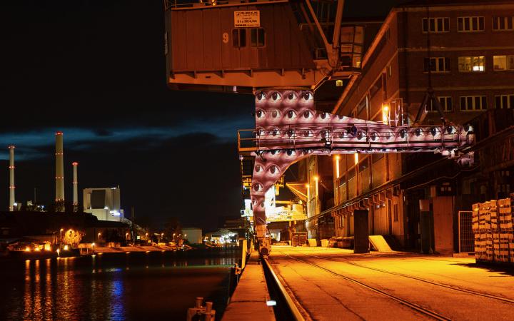 A port at night. Many pairs of eyes are projected on a column designed like an archway. The eyes look open in different directions.