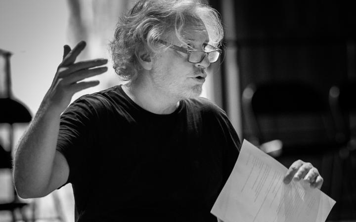 Black and white photo of Jean Pierre Seyvos, a man with curly hair and glasses, he is talking and holding a sheet of paper.