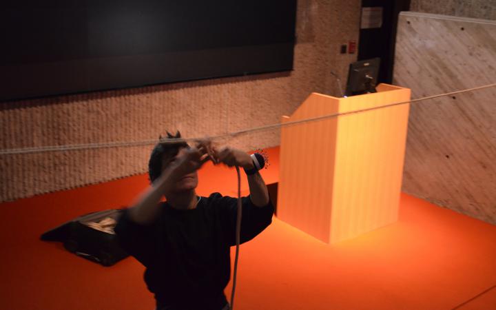 Das Foto zeigt eine Performerin im Vordergrund die an einem hängenden Seil ein weiteres befästigt. Sie trägt ein Nadelkissen um ihr linkes Handgelenk. Die schwarz gekleidete Person steht schräg vor dem Rednerpult auf dem orangenen Teppich.