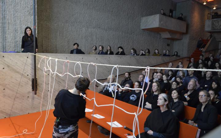 Foto eines gefüllten Vorlesungssaals. Die Künstlerin Judith Raum kreiert im Vordergrund ein Netz.