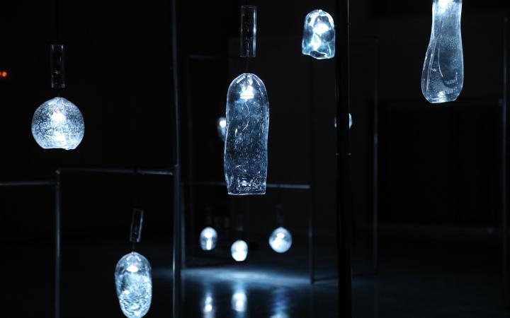 Glass vessels of various shapes hang in a dark room and glow dimly.