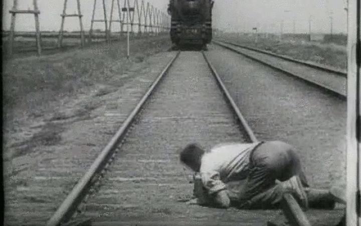 The black and white screenshot shows a track with a railway. A man lies crouching over a track and looks in the direction of the upcoming train.