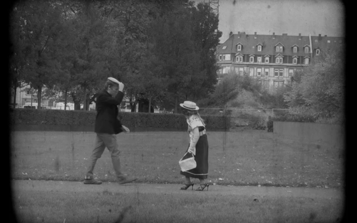 Two pedestrians meet on the street, the man is lifting his heat as to greet the Woman, that is aproaching. It is a still from a black-and-white movie made in the workshop "the little rascals". 